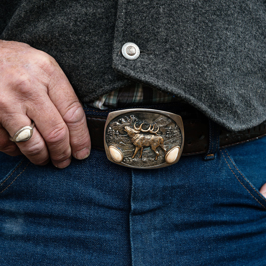 A man proudly wearing a sterling silver elk ivory belt buckle from Park City Jewelers, artistically designed with two prominently featured elk ivories, reflecting a stylish blend of personal hunting achievement and sophisticated craftsmanship.