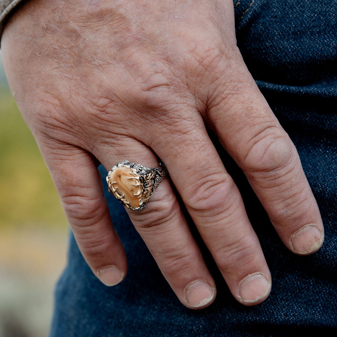 A man's hand sporting a distinctive two-toned gold and sterling silver elk ivory ring, featuring striking elk antler accents, exuding a sense of rugged elegance and a testament to his hunting heritage.