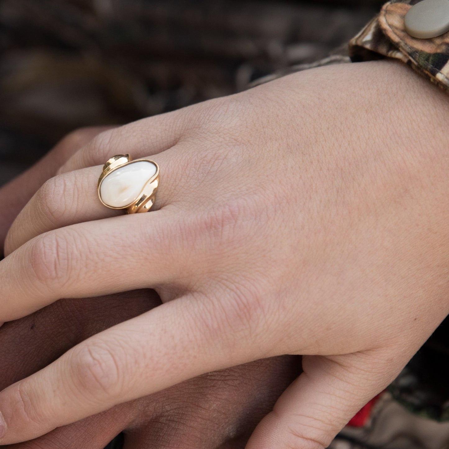 A woman's hand wearing the yellow gold elk ivory wave ring, resting on a man's hand with a camouflage sleeve in the background.