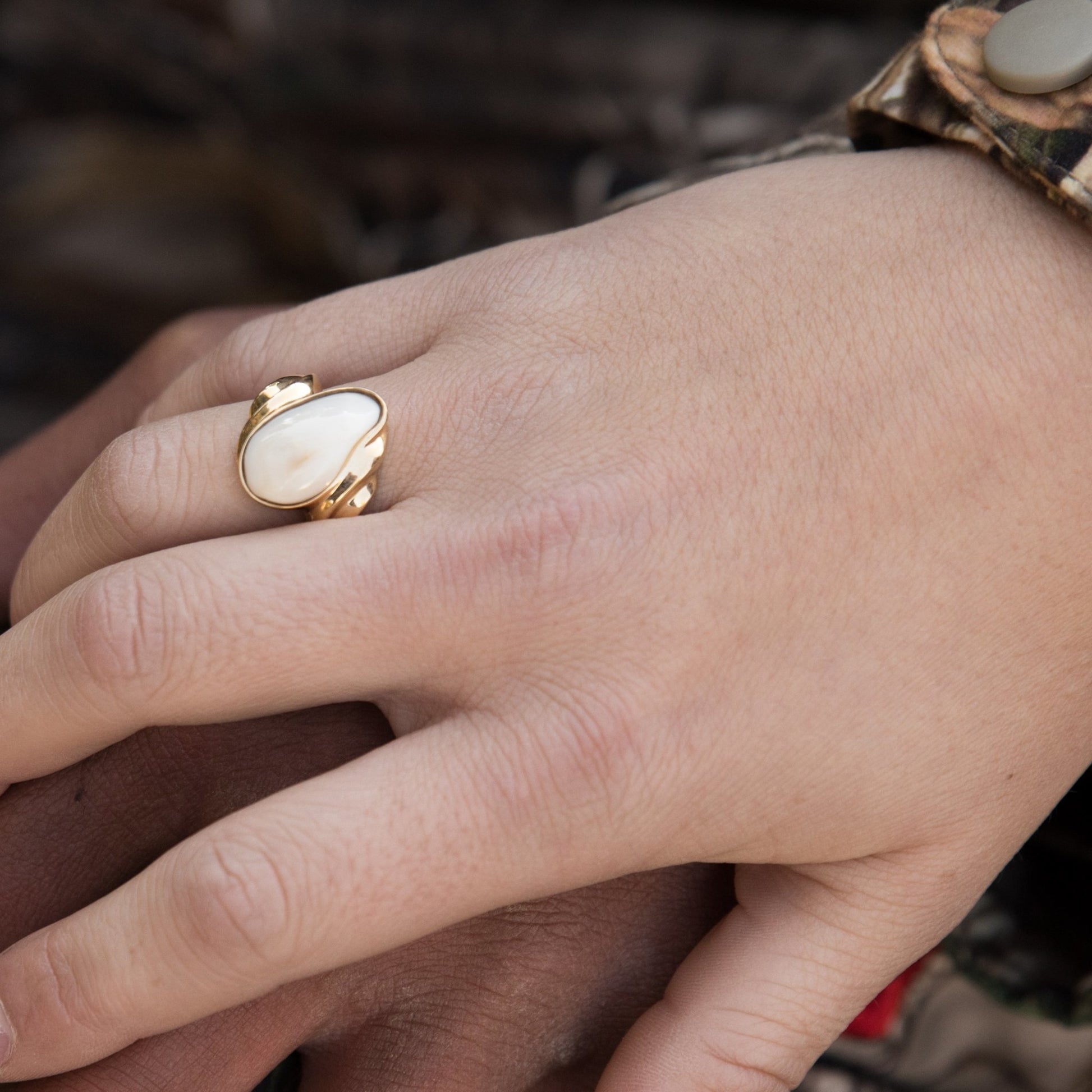 A woman's hand wearing the yellow gold elk ivory wave ring, resting on a man's hand with a camouflage sleeve in the background.